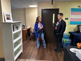 Councilmember Brianne K Nadeau speaks with HIPS staff member at an office with a bookshelf in the foreground the the trans pride flag hanging on the wall
