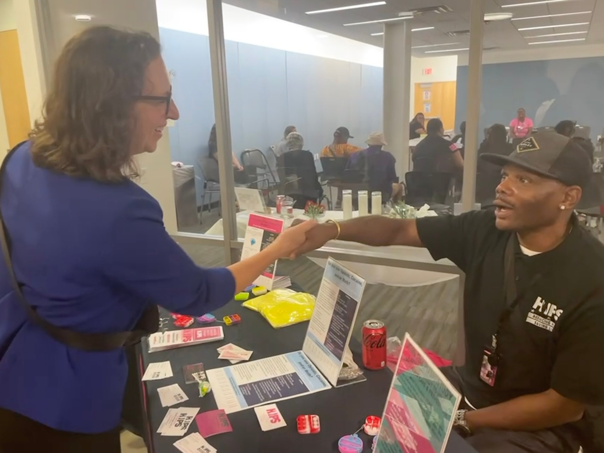 Councilmember Nadeau shakes hands with a man in a black HIPS polo shirt seated at a table with information pamphlets