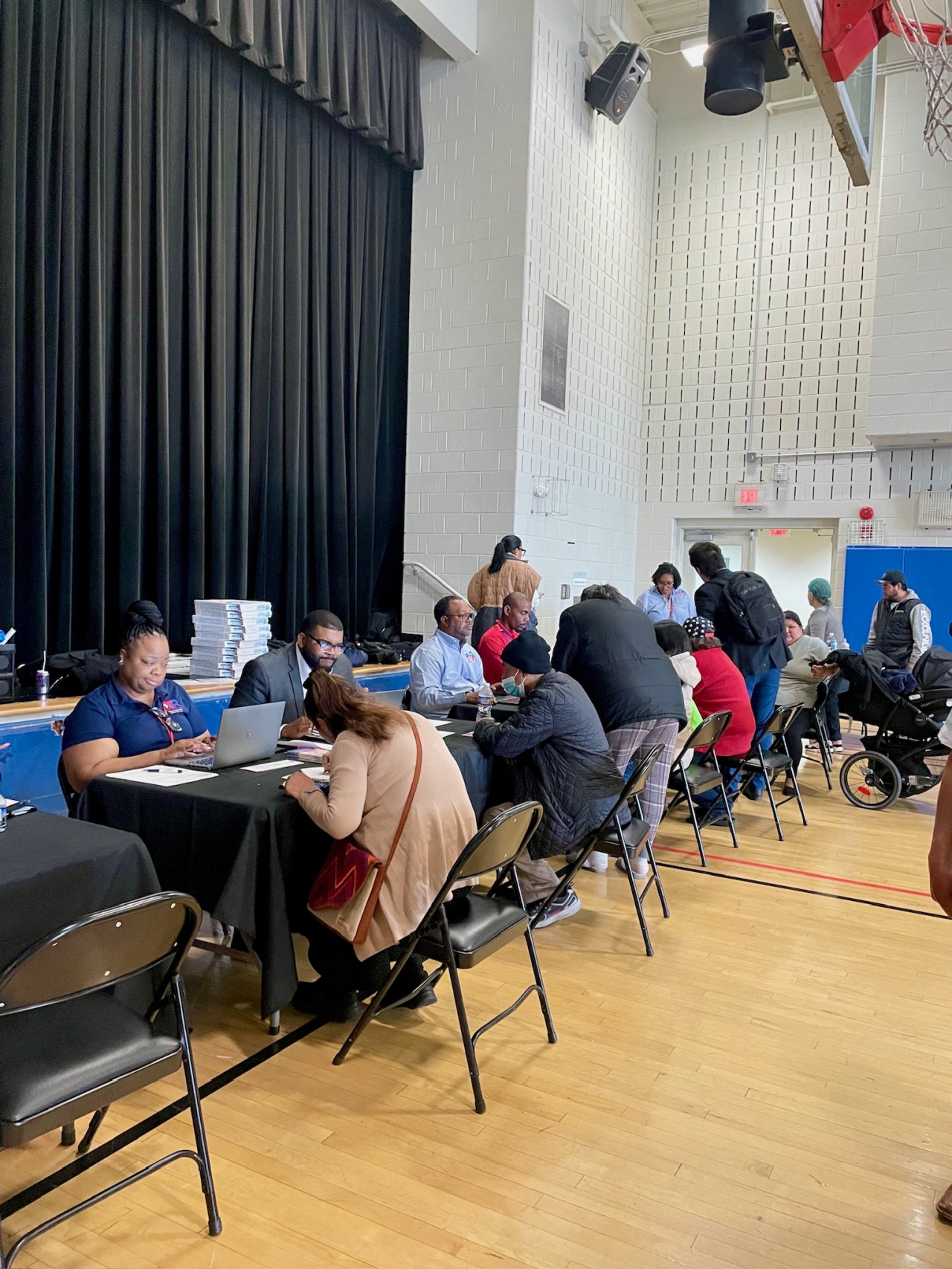 Workers are seated at a long table in a gymnasium with computers providing assistance to people seated across them.