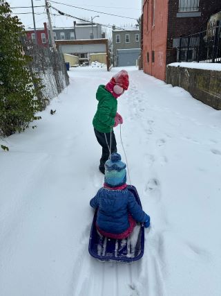 Two girls in winter coats and hats, one pulls a sled while another one rides in it down an alleyway in the snow