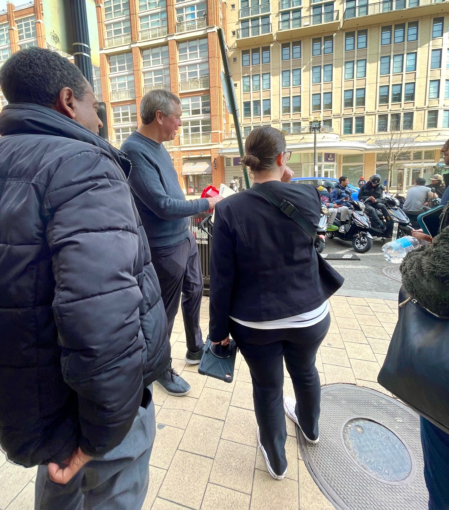 Councilmember Nadeau and several others stand on a sidewalk talking and pointing with Columbia Heights buildings in the background.