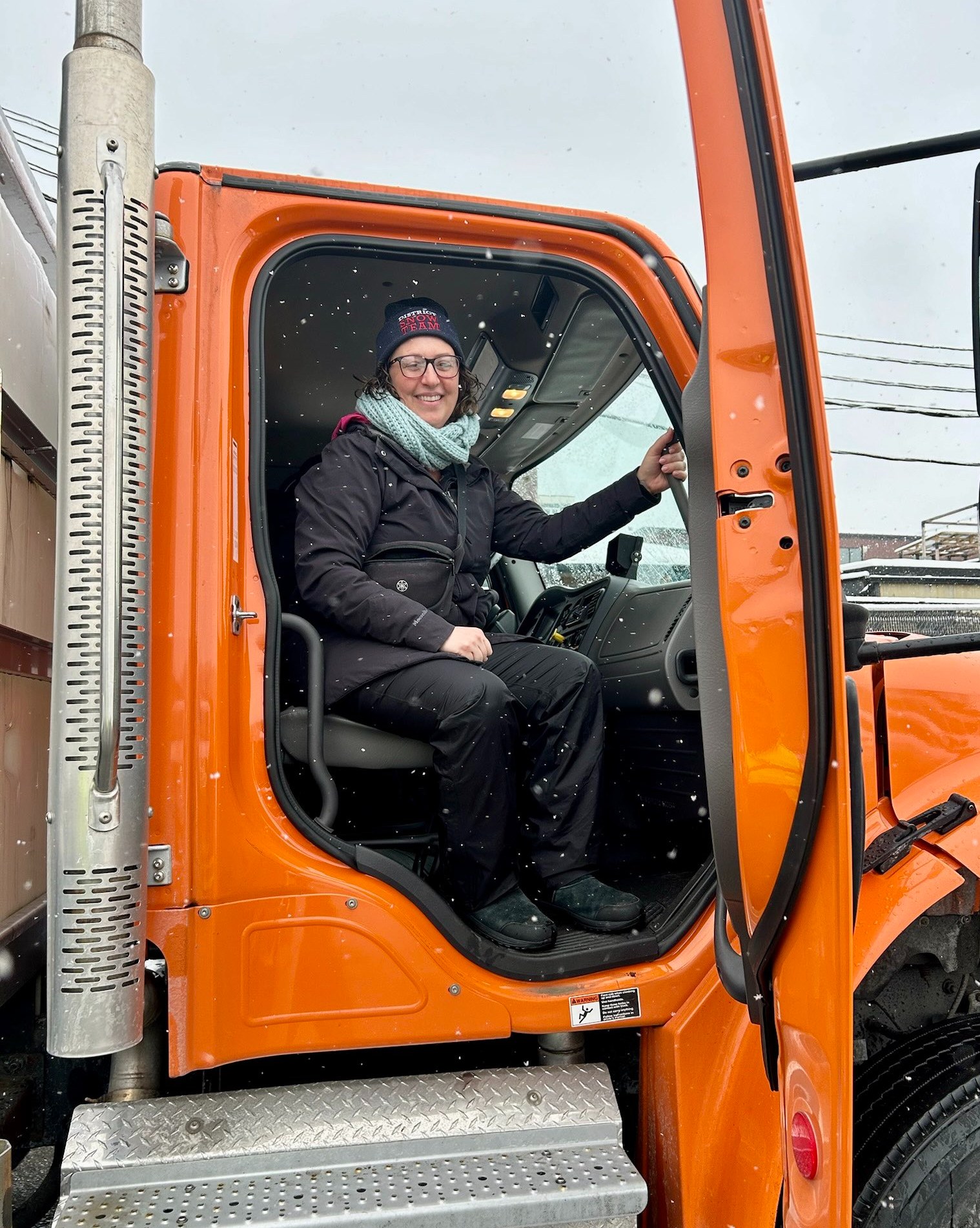Councilmember Nadeau sits in the passenger side of a parked snow plow, with the bright orange door opened, wearing a winter hat, scarf, winter coat, smilling broadly.
