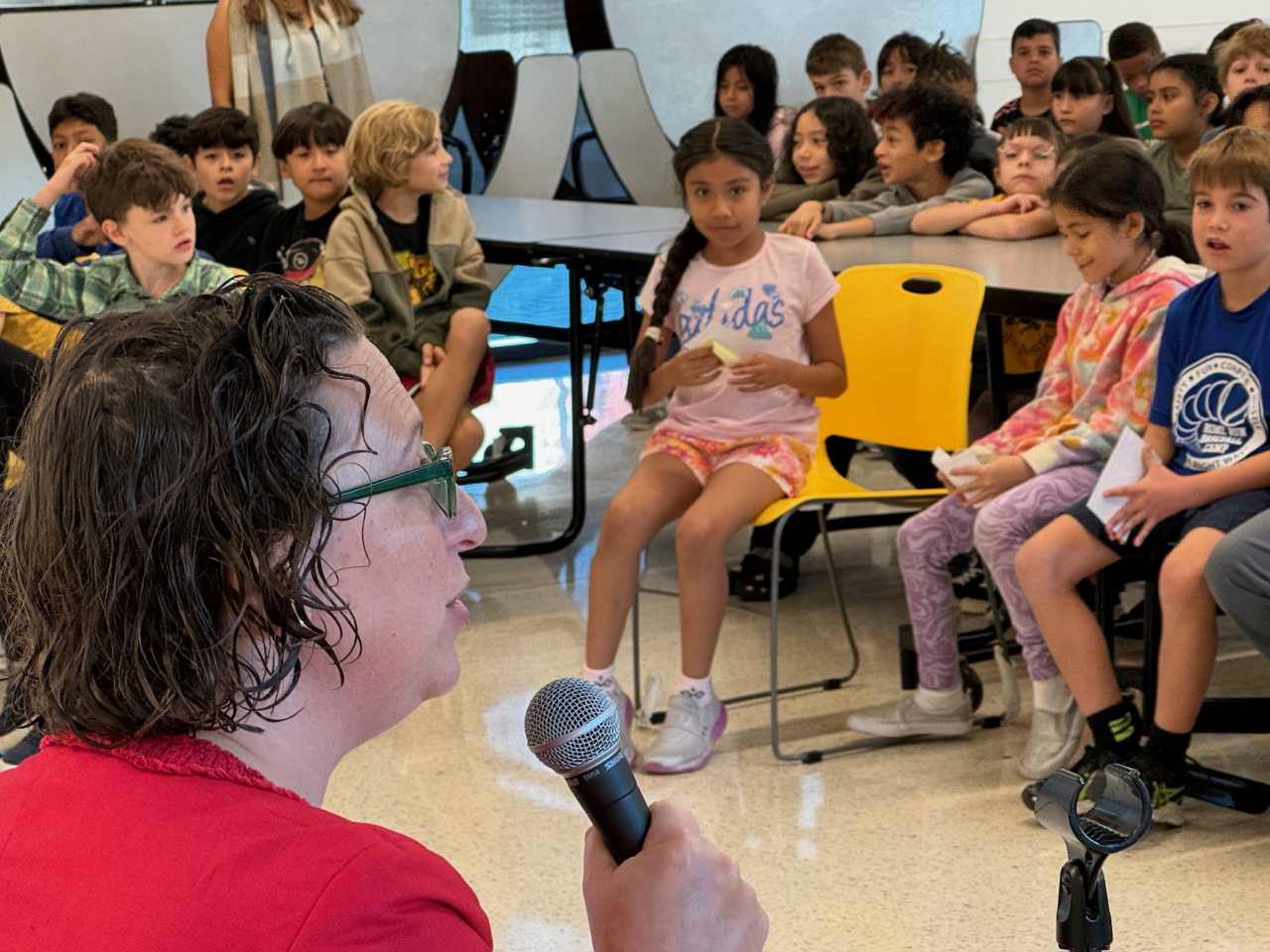 Councilmember Nadeau holds a microphone and speaks with a large group of third graders seated in a school cafeteria.