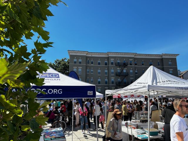 Multiple vendor tents on a plaza with many people milling about. An apartment building is in the background.