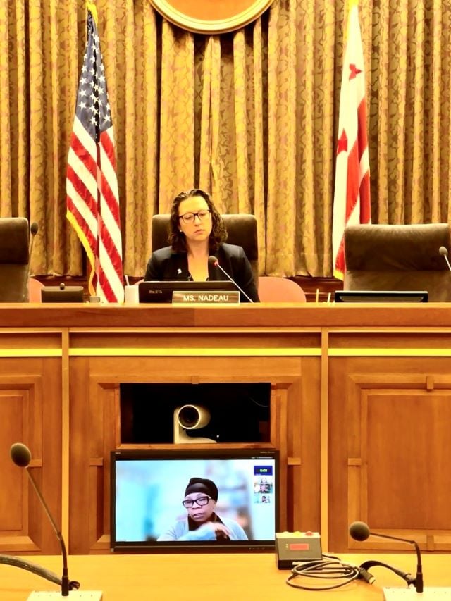 Councilmember Nadeau sits on the dais in the DC Council chamber while a witness appears via Zoom on the screen below. The US flag and DC flag are in the background.