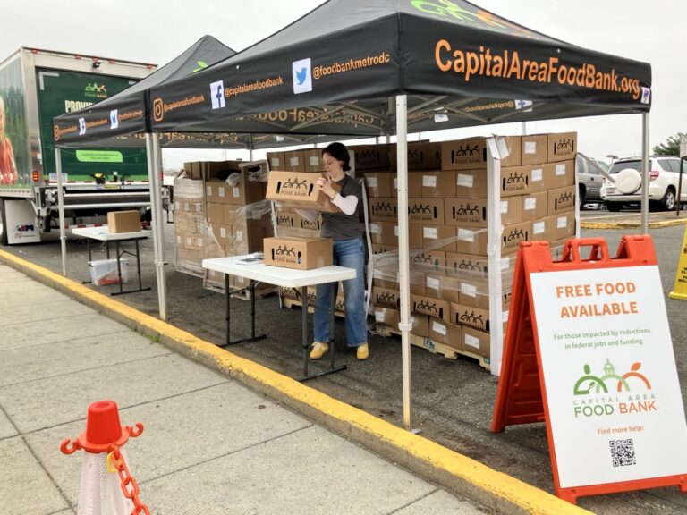 A woman stacks boxes of food provisions under an outdoor market-style tent. The words 
