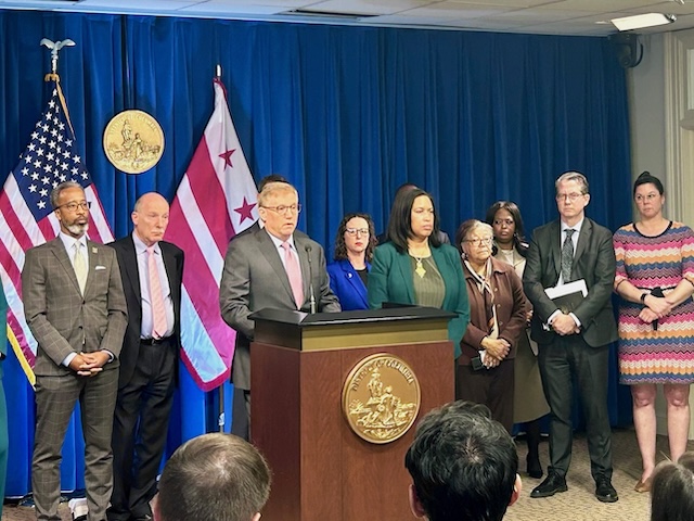 Councilmembers, the Mayor and the Chief Financial Officer stand behind a podium with the US and DC flags behind them and the city seal on the front of the podium.