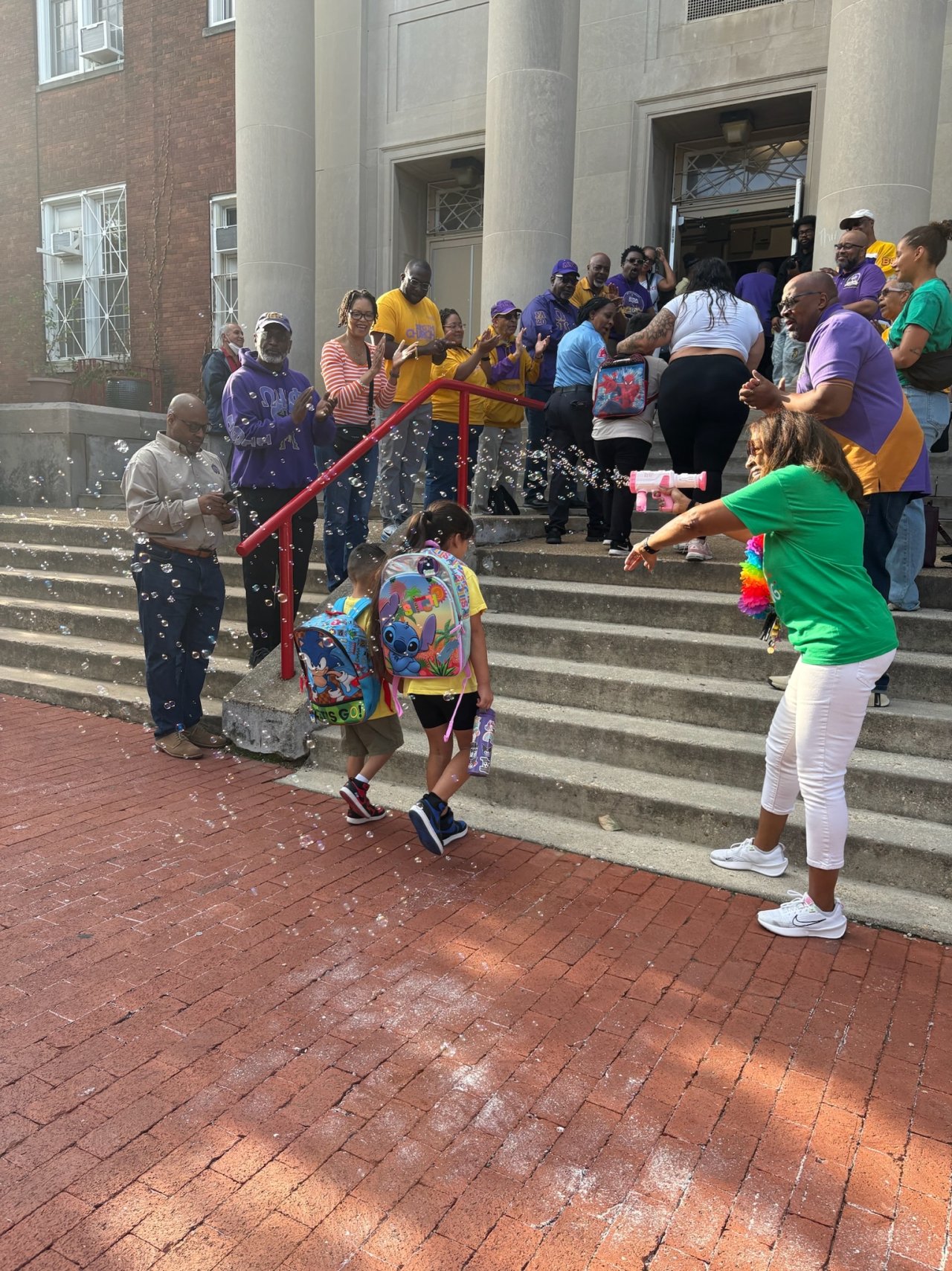 A crowd lines the steps on the way into a school as two students with backpacks walk up the steps