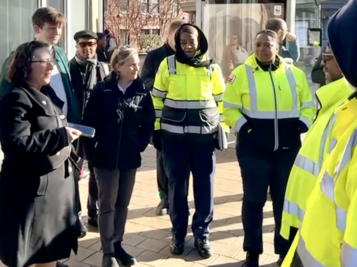 Group of 8-plus people, including three wearing bright yellow safety jackets, speaking on a sidewalk. Councilmember Nadeau is listening.