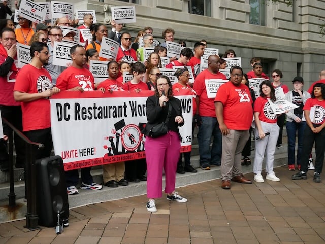 Councilmember Brianne K Nadeau speaks into a microphone in front of a crowd standing on the steps to the Wilson Building and wearing red t-shirts and holding a banner that reads 