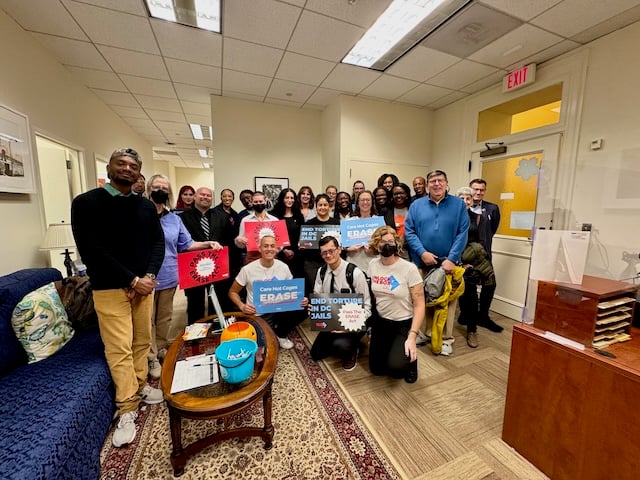 A group of people holding signs that say ERASE and end solitary confinement pose in an office lobby