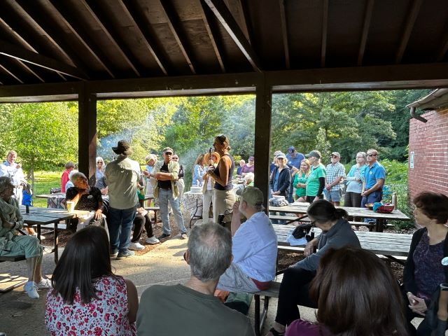 A dozen people are seated at tables under a covered picnic area, while another 20 or so are outside it. Smoke wafts upward from one or more grills.