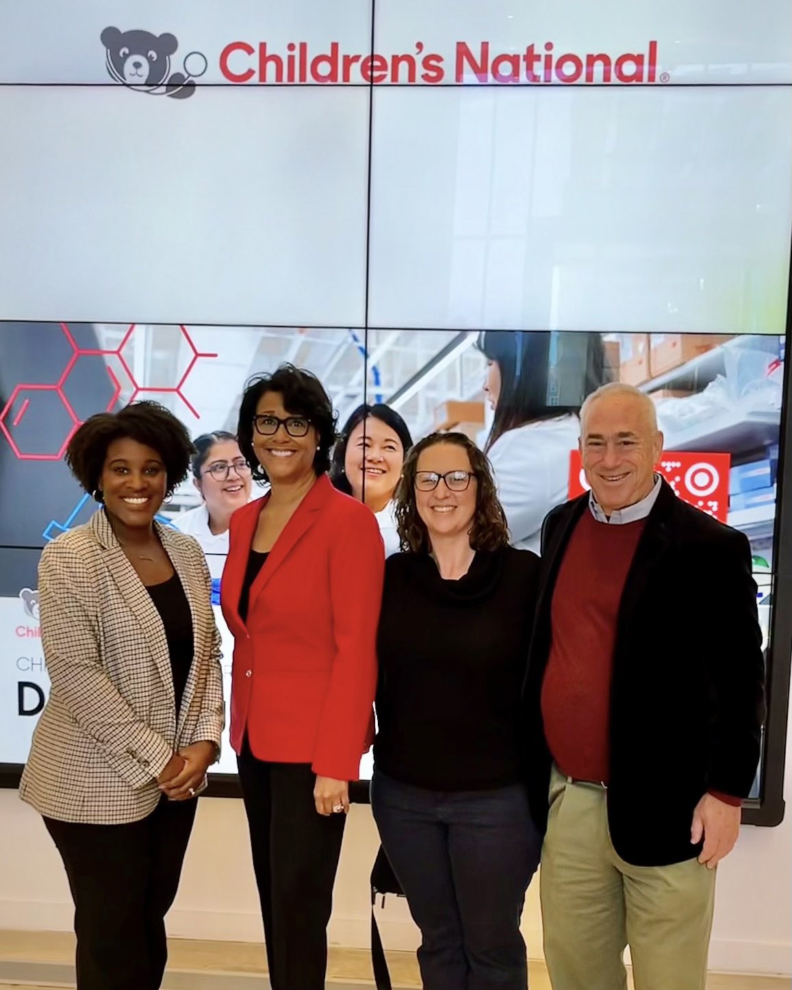 Three women and one man stand in front of digital sign reading 