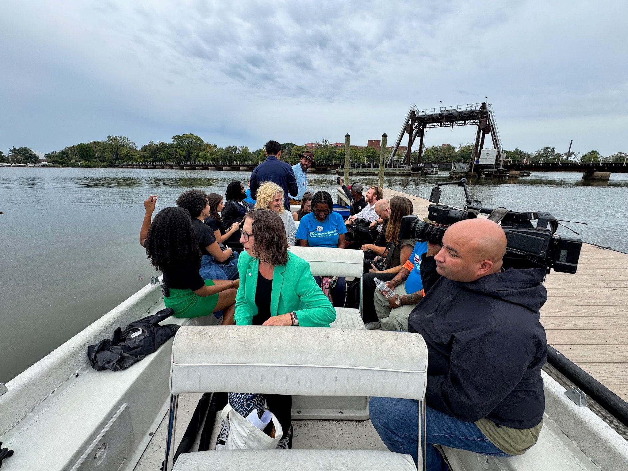 Photo of people in boat docked in Anacostia River. Councilmember Brianne K Nadeau wears a green jacket and is seated next to a man holding a TV camera.