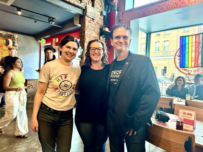 Councilmember Nadeau and Leah from Roght Proper and Johnny from HIPS pose for a photo with other event participants milling in the background. A storefront window in the background looks out into the street with a building in the background.
