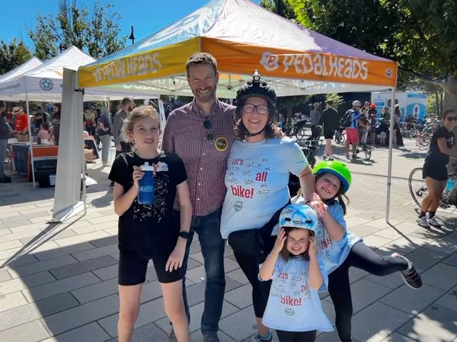 Councilmember Nadeau with Councilmember Allen and their children in front of a Pedalheads tent on a stone plaza.