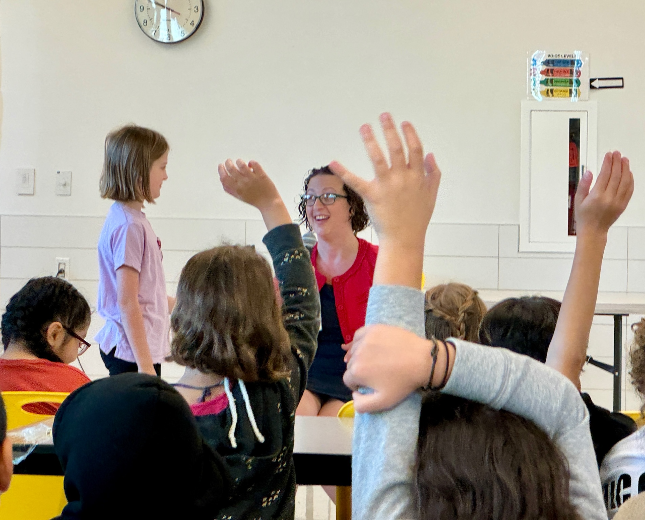 Backs of kids heads with their hands raised while seated in cafeteria seats. Councilmember Nadeau is visible smiling, speaking to a young student standing in front of her.