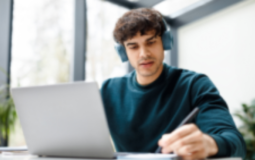 A person wearing headphones, with an open laptop in front of them and holding a pen.