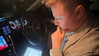 Paul Slevin sitting in the drivers seat of a car and looking at OU course pages on a tablet.